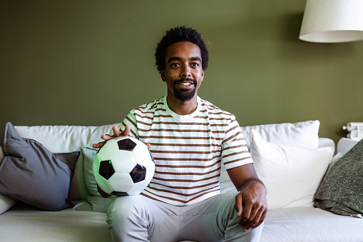 A young Afro American man embraces his love for soccer at home. Sitting on the sofa with a soccer ball in hand, he excitedly watches a match on TV.