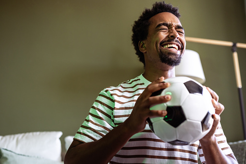 With adrenaline running high, an excited Afro American man sits on the sofa with a soccer ball in hand, fully engaged in the soccer spectacle on TV.