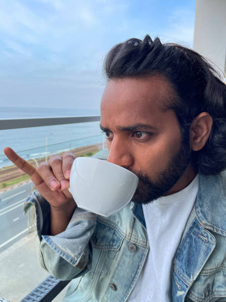 Full frame image of Indian man on hotel balcony with glass safety barrier, drinking coffee from white mug, extended little finger, Coastal (southern) railway line parallel to highway and coastline, sea background, Colombo, Sri Lanka Stock photo showing close-up view of man sitting on hotel balcony in sunshine with a background view of railway train tracks of Coastal (southern) railway line and sea in Colombo, Sri Lanka. pinky finger stock pictures, royalty-free photos & images