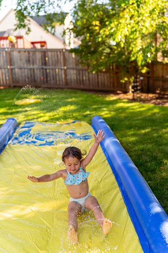 Preschool age girl Eurasian descent smiles as she slides down a Slip N' Slide with her hands raised up in the air. The toddler is having fun playing in the water on a hot summer day.