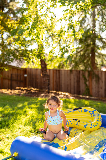Preschool age Eurasian girl  sits on a Slip N' Slide in the backyard of her home and smiles while playing outside on a hot, summer day.