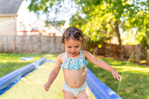 An adorable two year old Eurasian girl giggles while running through water spraying from a slip n slide in the backyard of her home on a hot summer day.