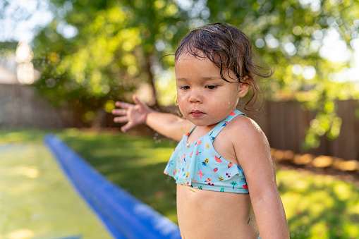 A cute Eurasian toddler girl reaches her hand towards water spraying from a backyard Slip N' Slide while playing outside at home on a warm, summer day.