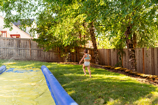 Wide shot of an excited two year old Eurasian girl playfully running away from the camera while playing on a Slip N' Slide in the backyard of her home on a hot, summer day.