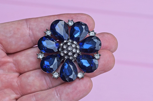 one round silver jewelry brooch in the shape of a flower with white and blue stones lies on the fingers on the hand on a pink background