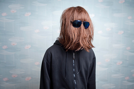 A man's long red hair hair completely cover his face with the exception of his glasses.  Vintage retro wallpaper in the background.  Horizontal with copy space.