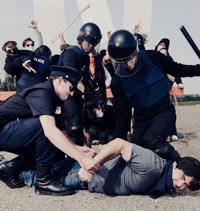 Image of a protest taking place and a male being arrested by a group of police man. Protesters are in the background with blank signs yelling at the officers.