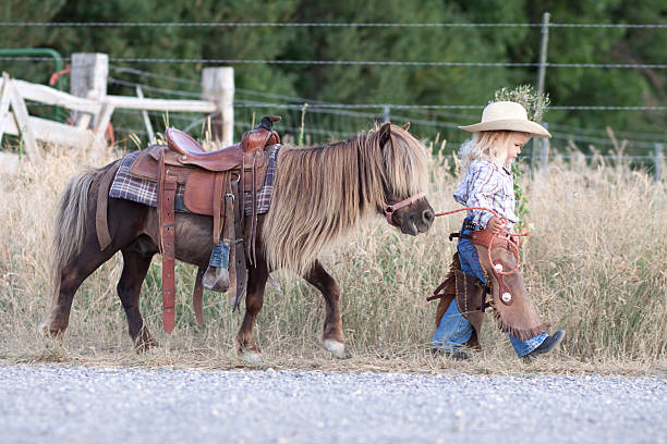 Small child in cowboy outfit with cute hairy pony on a lead Little girl with cowboy hat, walking with her little horse. pony photos stock pictures, royalty-free photos & images