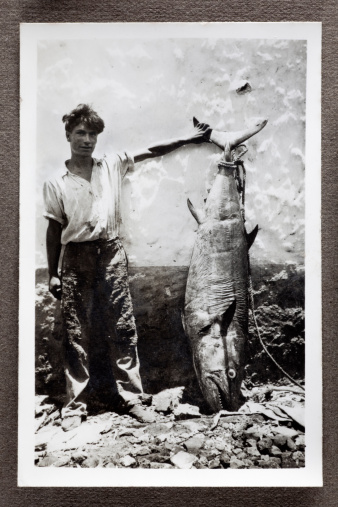 Vintage black and white photograph of a peasant fisherman proudly showing off a large Bluefin Tuna leaning against a wall. Some dust and scratches which convey age of original image, taken in Mauritius, 1942.