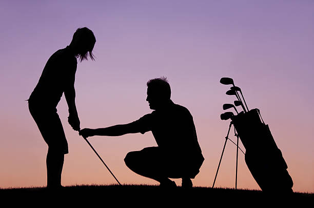 A silhouette of a woman getting a golf lesson from a golf professional and instructor. Themes of the image include golf lessons, instruction, short game, sport, game, practice, teaching, learning, lesson tee, driving range, golf professional, chipping, evening, model, young, woman, attractive, side view, 30s, helping, assisting, and golf academy. 
