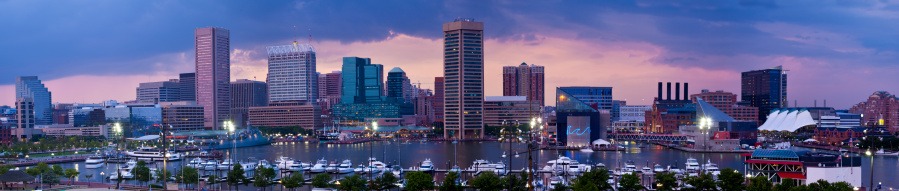 Panorama of Baltimore's Inner Harbor at dusk with thunderstorms approaching. Numerous buildings and skyscrapers dot the skyline.