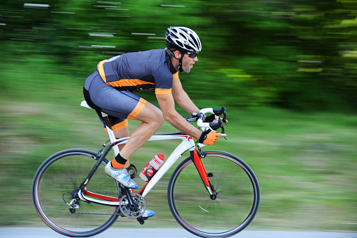 Recreational male and female cyclists riding the bicycles so fast the background is blurred. The couple is located in the countryside on an empty road surrounded by green trees. The couple is wearing professional cycling gear and helmets. Couple having an active weekend.