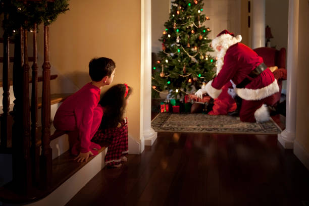 Peeking at Santa Two children peek at Santa Claus placing gifts under the Christmas tree. santa stairs stock pictures, royalty-free photos & images