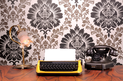 Typewriter on a desk with lamp and phone. All materials are  old and photographed in front of a nostalgic wallpaper.