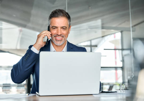 istock Happy busy middle aged business man talking on phone using laptop in office. 1575126019