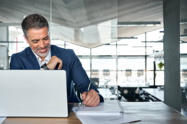 istock Busy older business man working on laptop in office at desk writing notes. 1575126029