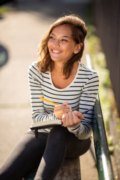 istock smiling young woman sitting on bench with pen and book 1575128612