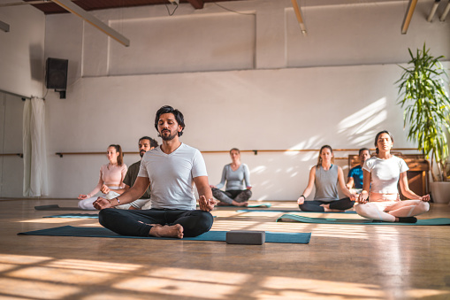 Low angle shot of a mid adult, focused male Indian man sitting in lotus pose and meditating in a session with other people behind him. Full length image.