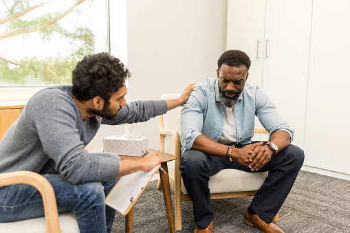 The young adult male therapist comforts his distressed mid adult male client by placing his hand on the man's shoulder.