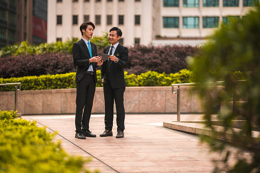 Full length shot of two asian businessmen having an outdoor meeting. They are both wearing formal outfits and discuss work using a digital tablet.