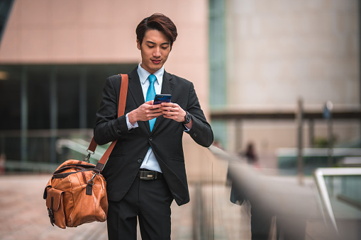 Short getaway trip. Young chinese businessman checking his cellphone while holding a big brown travel bag. He is wearing a black suit and checking his upcoming itinerary.