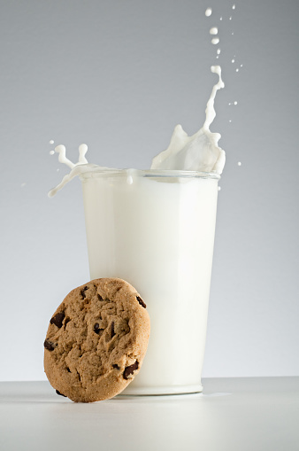 A glass of splashing milk with a chocolate chip cookie leaning on the glass. 
