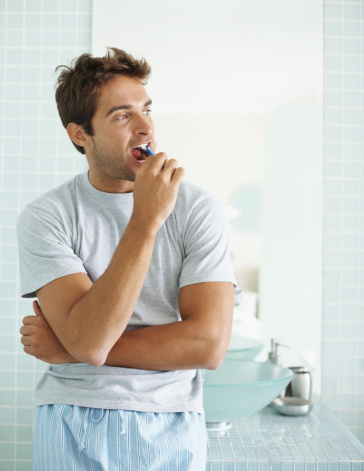 Young man brushing his teeth in the bathroom looking away in thought