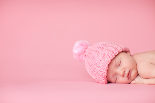 Color photo of a beautiful newborn baby girl wearing a knit hat while sleeping peacefully on a pink background. Composed with room for text on left.