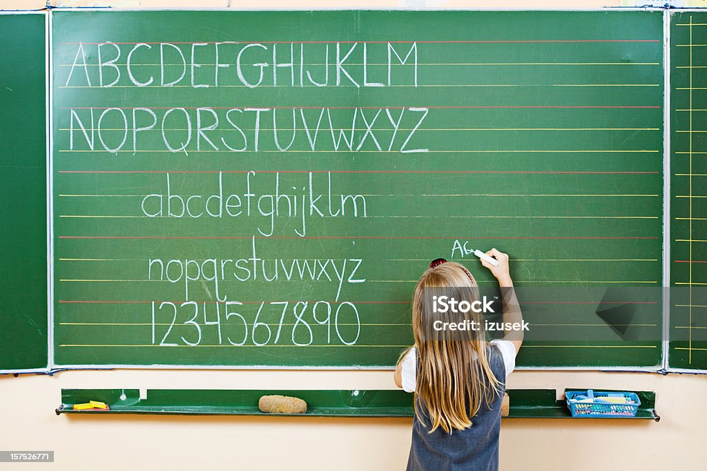 School Girl Writing On Blackboard, Rear View A school girl writing on a blackboard with alphabet and numbers written on it. Chalkboard - Visual Aid Stock Photo
