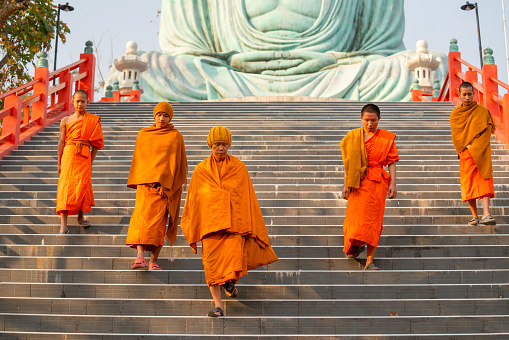 Group of young and senior monks walk down from area of big buddha statue with day light.