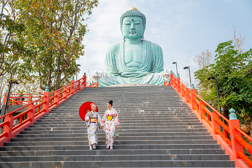 Two Asian young woman with japanese style dress walk down on stair with green big buddha statue in background with day light.