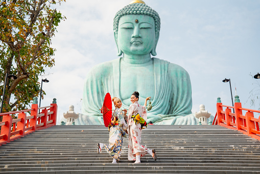 Two Asian young woman with japanese style dress stay on stair in front of green big buddha statue with day light and they look happy.
