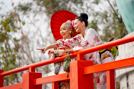 Two Asian pretty women stay close to red railing in the area of base of green big buddha statue and one also point to left side and they look happy with smiling.