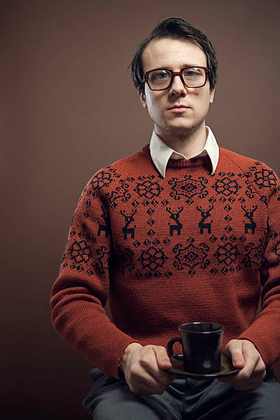 Vintage Nerd With Reindeer Sweater A nerdy young man with big glasses and a cool retro reindeer sweater looks at the camera with a serious calm expression, holding a cup of coffee or tea with a saucer.   Shot indoors on a vertical brown background with copy space. nerd sweater stock pictures, royalty-free photos & images