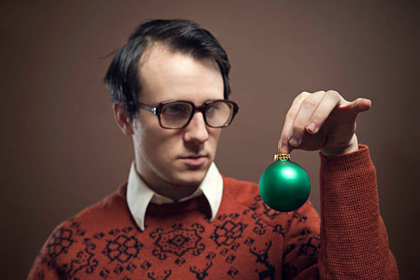 Vintage Nerd With Reindeer  Sweater It's Christmas time, and a nerdy young man wearing big glasses and a retro sweater contemplates where to hang his green bauble ornament.  Shot indoors on a horizontal brown background with copy space. vintage nerd with reindeer sweater stock pictures, royalty-free photos & images