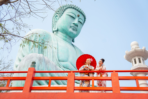 Wide shot of two Asian pretty women stay close to red railing in the area of base of green big buddha statue and they look to left side with happiness.