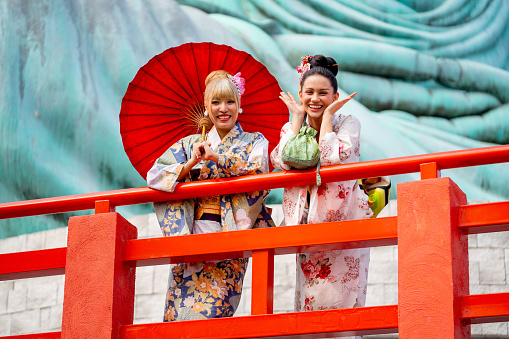 Two Asian pretty women stay close to red railing in the area of base of green big buddha statue also look at camera with smiling.