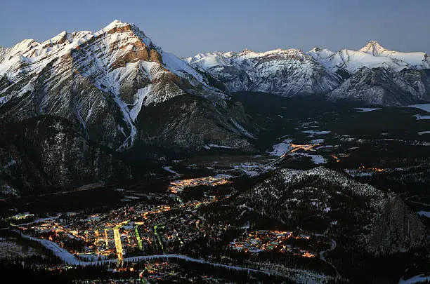 An aerial view of Banff, Alberta from Sulphur Mountain. This long exposure and night image of the tourist town of Banff highlights the beauty of the Canadian Rockies. Snow is covering the nearby mountains. Closet mountain is Cascade Mountain with Fairholm Range in the distance. The bright street is Banff Avenue where many of the hotels, restaurants, and shops are located. Banff is located approximately two hours west of Calgary. 