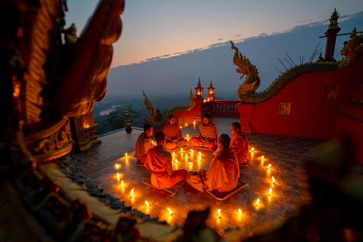 Side view of young and senior monk sit in a circle with several lighting candle around in area of front of chapel or church for meditation in early morning with blue dark sky.