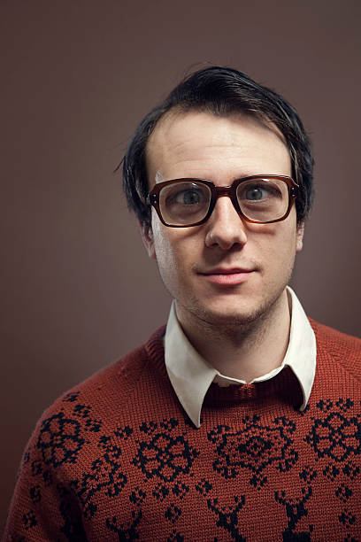 Vintage Nerd With Reindeer Sweater A nerdy young man with big glasses and a cool retro reindeer sweater looks at the camera with a serious calm look of contentment.   Shot indoors on a vertical brown background with copy space. vintage nerd with reindeer sweater stock pictures, royalty-free photos & images