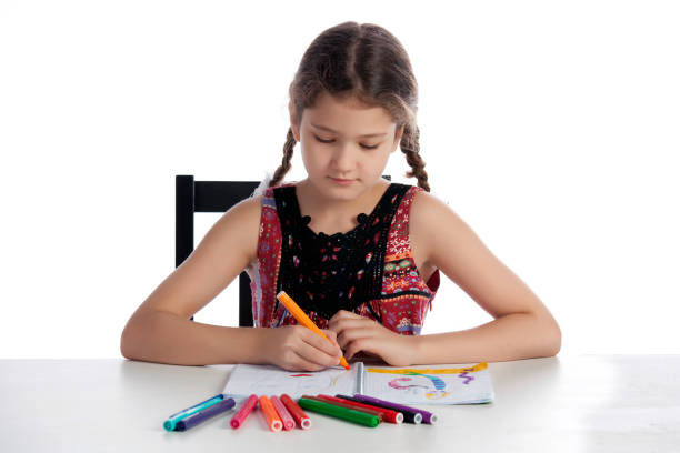 Girl sitting at a white table and drawing with colored felt brushes in notebook. Isolated on white background