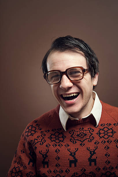 Vintage Nerd With Reindeer Sweater A nerdy young man with big glasses and a cool retro reindeer sweater laughs loudy with a big toothy smile on his face.   Shot indoors on a vertical brown background with copy space. vintage nerd with reindeer sweater stock pictures, royalty-free photos & images