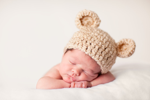 Color photo of a newborn baby wearing a knit hat with bear ears while sleeping peacefully on a soft blanket.