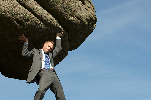 Businessman struggling with lifting a massive boulder over his shoulders outdoors in blue sky