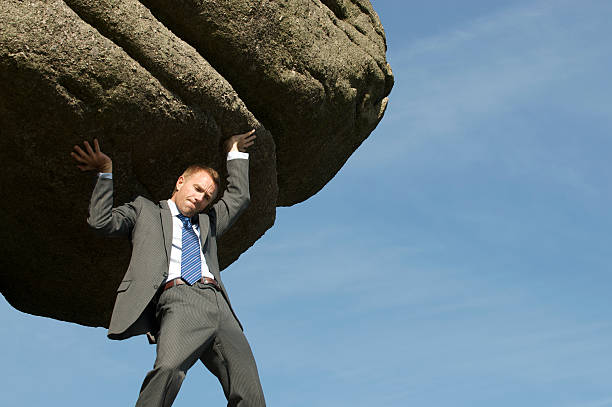 Businessman Struggling Lifting Massive Boulder Outdoors in Sky Businessman struggling with lifting a massive boulder over his shoulders outdoors in blue sky sisyphus stock pictures, royalty-free photos & images