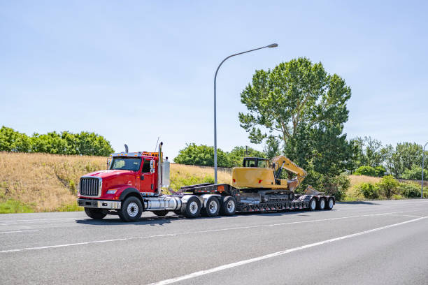 istock Red day cab powerful big rig semi truck tractor transporting excavator on step down semi trailer driving on the highway road exit 1575340735