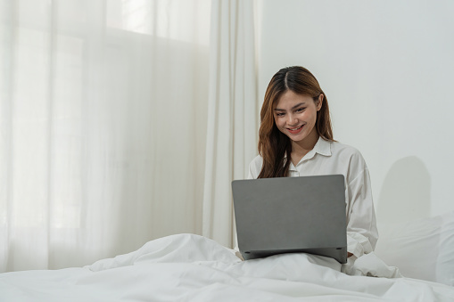 Beautiful happy asian woman lying in bed relaxed and looking at laptop screen. Girl smiling happy while browsing internet on computer, resting at home.
