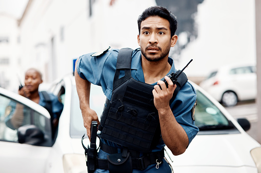 Asian man, police and walkie talkie with gun in city for suspect, communication or reinforcements. Serious male person, security guard or cop radio calling backup for crime in street of an urban town