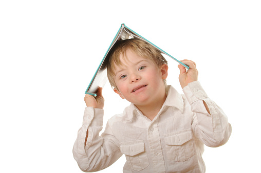 Boy loves reading. Seven year old boy with Down Syndrome dressed in white clothing with a turquoise book on his head. White background.