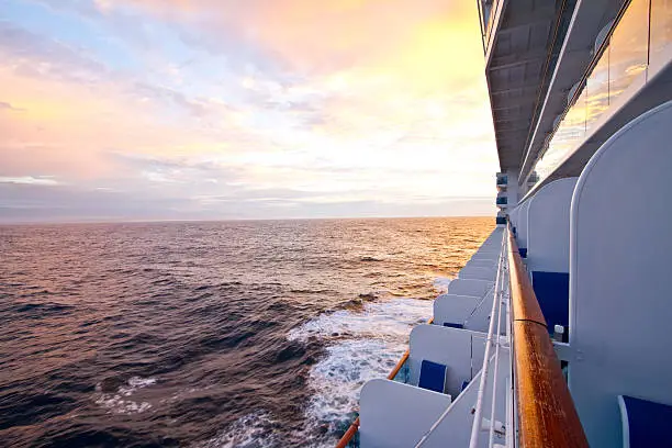 Shot along the side of a large cruise ship in the Caribbean sea from a balcony suite.  Image looks toward the back of the ship showing the wake under a brilliant cloudscape sky at sunrise or dawn.